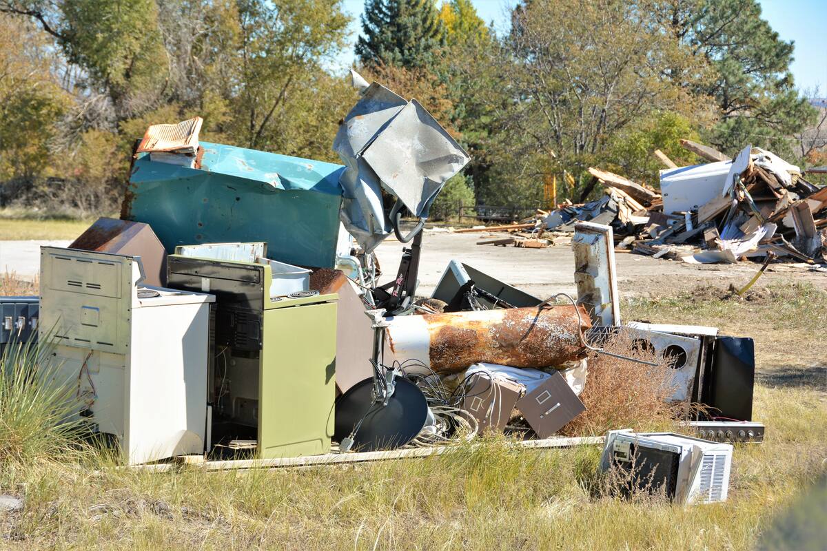 A heap of old appliances and scrap: a white electric range, an avocado-green range, a rusted water heater, a window AC unit, a satellite dish, an exercise machine, sheet metal and cable