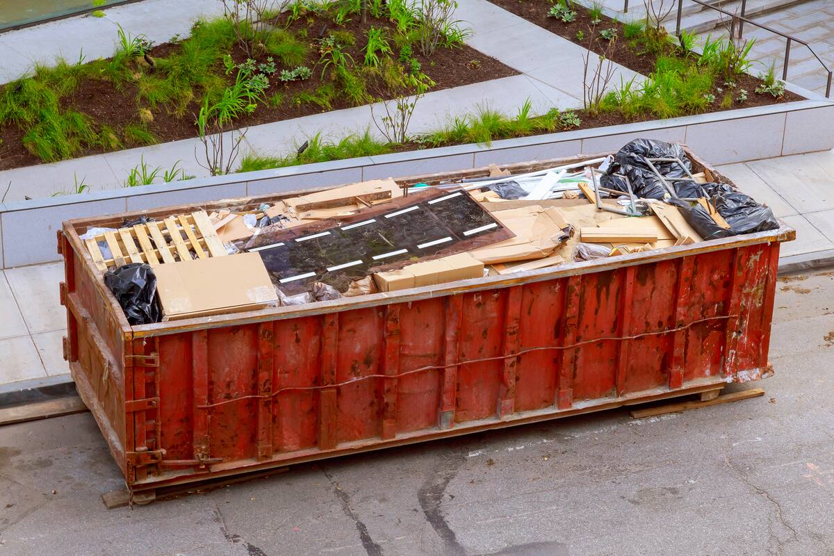 A full red roll-off container of a wooden pallet, flattened cardboard and boxes, scrap boards, a dark glass panel, metal pipes and black trash bags